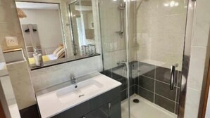 Ein modernes Badezimmer mit einer Duschkabine aus Glas, einem weißen Waschbecken, dunklen Schränken und einem Spiegel über dem Waschbecken.