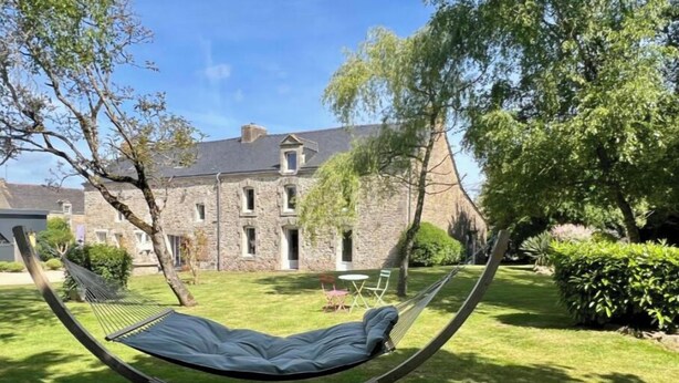 Eine Hängematte in einem grünen Garten, im Hintergrund ein Steinhaus.