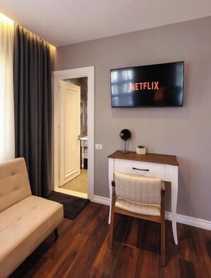 Una sala de estar con un sofá color beige, una mesa de centro de madera, un televisor montado en la pared mostrando 'NETFLIX' y una puerta que da a otra habitación.