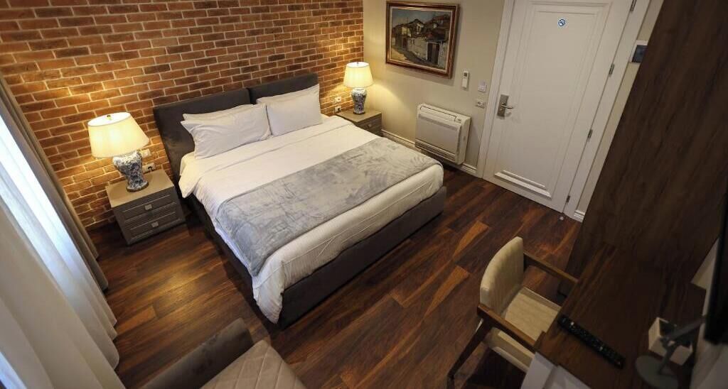 Une chambre d’hôtel avec un mur en briques, un lit avec du linge de maison blanc, deux tables de chevet avec des lampes, un radiateur et une porte.