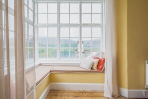 Ein Zimmer mit einem großen Erkerfenster, einer Holzbank und Blick auf einen See und Berge.