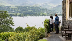 Una pareja contemplando un lago con barcos, rodeada de vegetación y montañas.