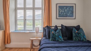 Un dormitorio con un ventanal grande, una cama con muchas almohadas, una mesita de noche con una lámpara y un cuadro en la pared.