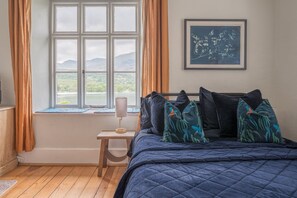 Ein Schlafzimmer mit großem Fenster, einem Bett mit vielen Kissen, einem kleinen Tisch mit Lampe und einem gerahmten Bild an der Wand.