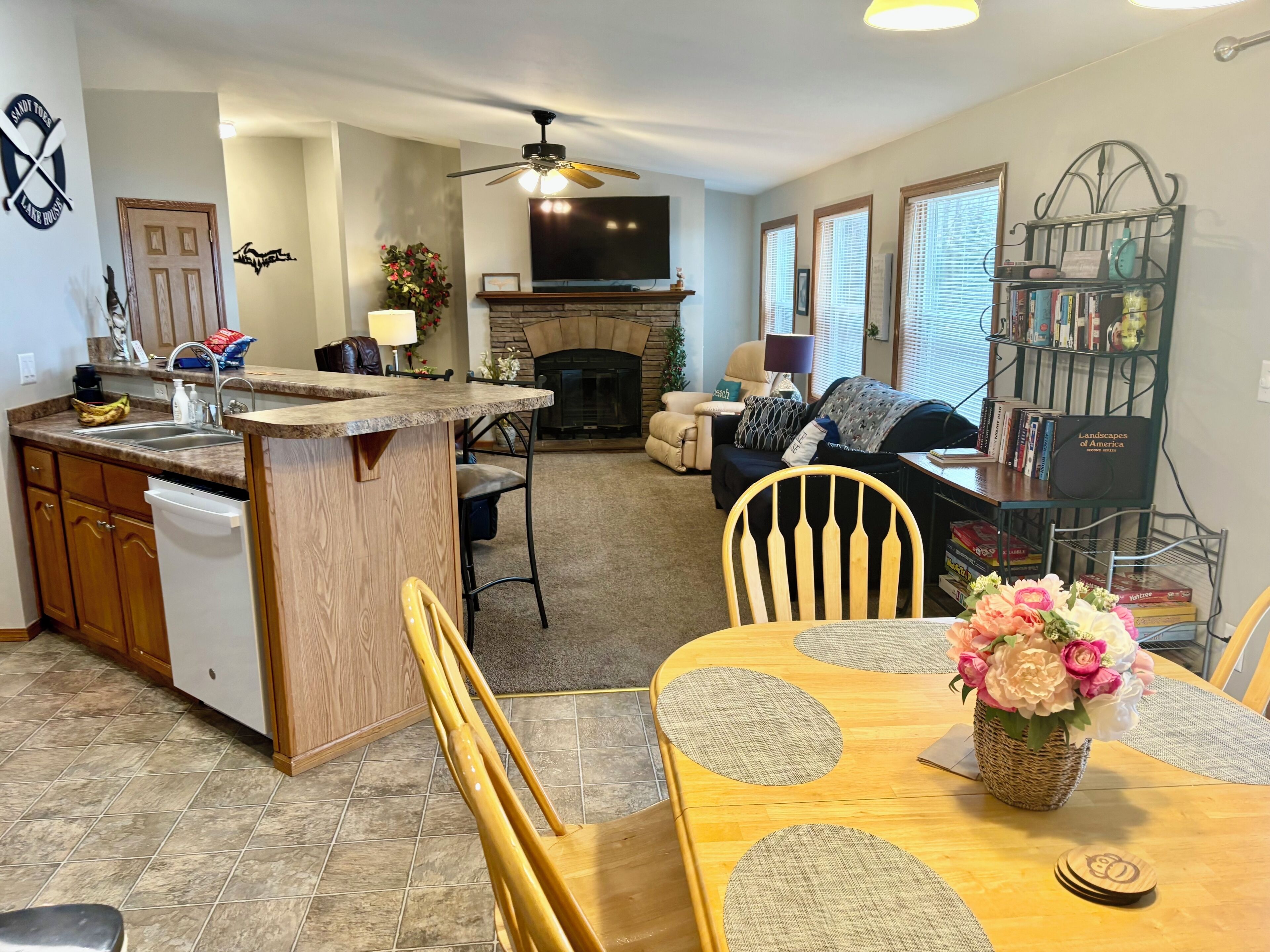 A modern kitchen with a built-in island, a dining area with a wooden table and chairs, a living room with a sofa and a fireplace, and a bookshelf filled with books.