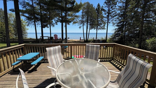 A deck with a glass table and striped chairs, overlooking a beach with trees and a clear sky. - Sandy Toes Retreat,stunning view & just 25 steps to a sandy beach Lake Michigan. (Cedar River)