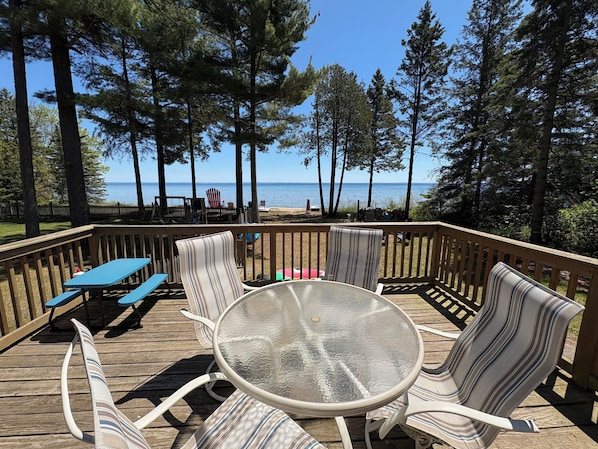 A deck with a glass table and striped chairs, overlooking a beach with trees and a clear sky.