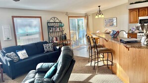 A living room with a sofa, a coffee table, a bookshelf, and a kitchen area with a counter and bar stools. - Sandy Toes Retreat,stunning view & just 25 steps to a sandy beach Lake Michigan. (Cedar River)