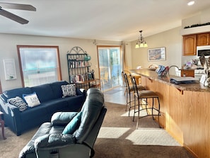 A living room with a sofa, a coffee table, a bookshelf, and a kitchen area with a counter and bar stools.