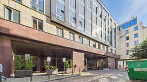 Hotel moderno com entrada coberta, pátio pavimentado e contentor de lixo verde.