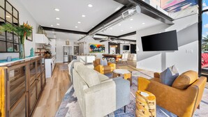 A modern interior with a kitchen, dining area, and living room, featuring wooden floors, a large sofa, and a flat-screen TV. - Sleeps 10 in Jax Beach - 2BR/2BA - Pool Table + Firepit (Jacksonville Beach)
