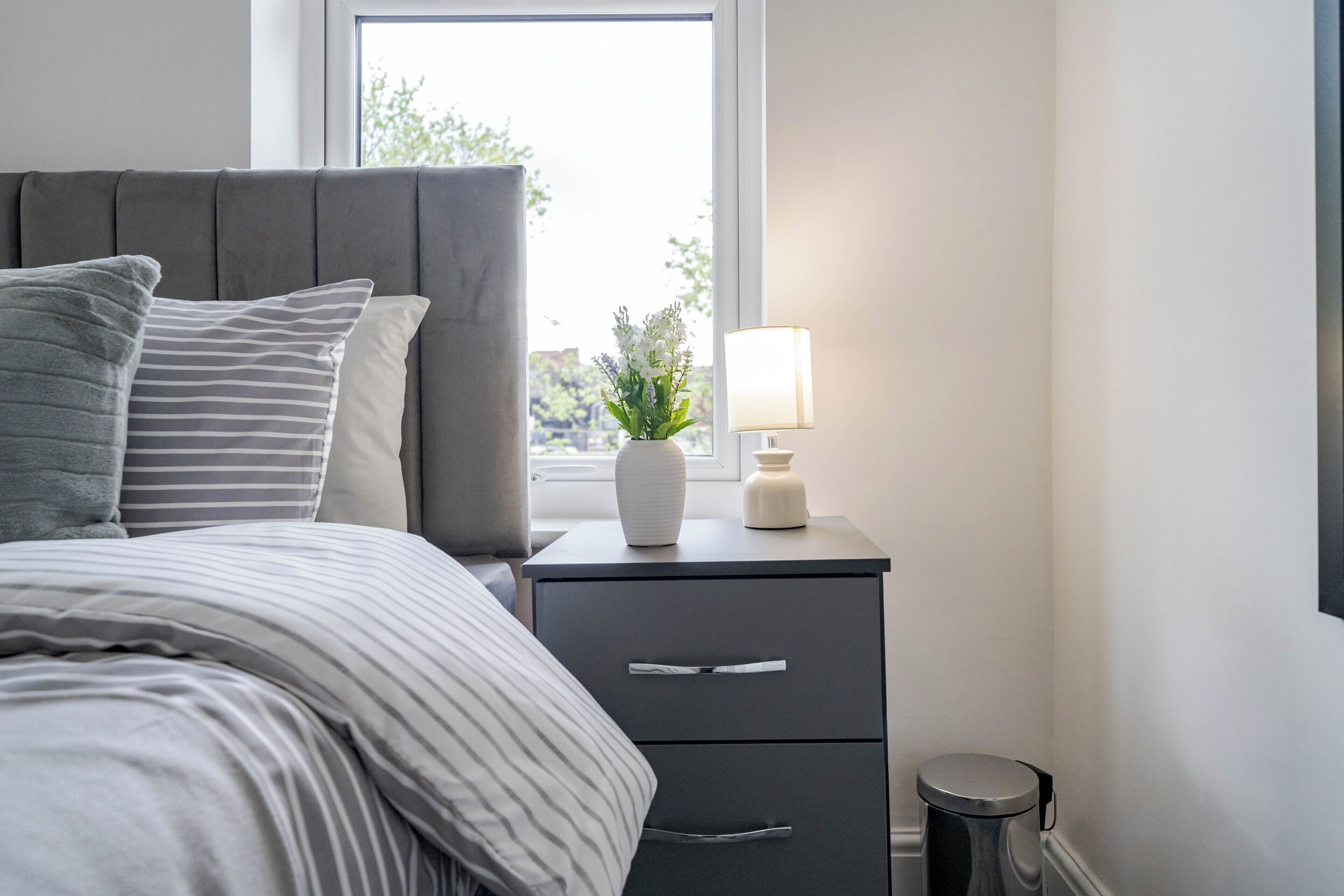 A neatly made bed with striped bedding, a bedside table with a lamp, a small plant, and a trash bin.