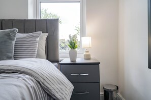 Een netjes opgemaakt bed met streepjesbeddengoed, een nachtkastje met een lamp, een kleine plant en een prullenbak.