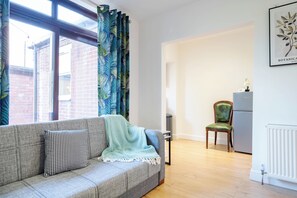 Een woonkamer met een grijze bank, een gekleurd kussen, een groene plaid, een stoel, een radiator en een raam met gordijnen.