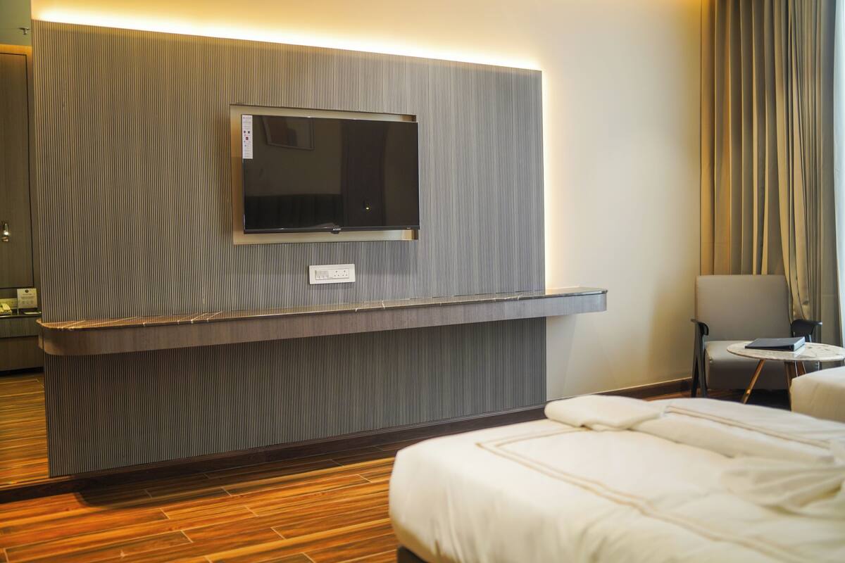 Una habitación de hotel con una televisión de pantalla plana en una pared texturizada, una cama con ropa blanca, una mesita redonda con una silla y una ventana con cortinas.