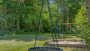 Ein Spielplatz mit einer Taitschaukel, einer Schaukel und einem Sandkasten in einer bewaldeten Gegend.