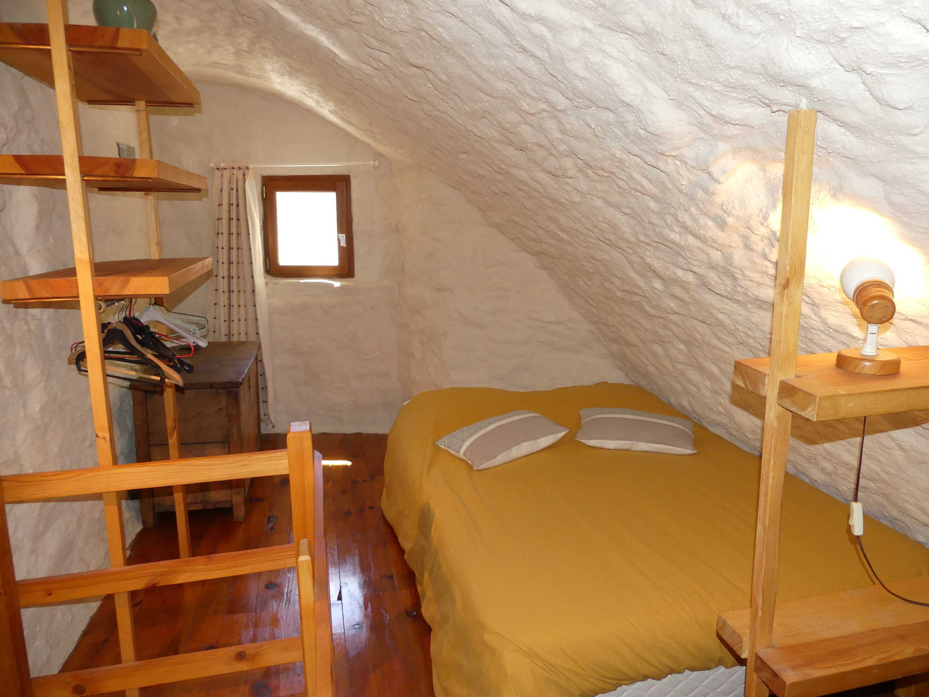 Um quarto no sótão com escada de madeira, uma cama com almofadas, uma pequena janela e um abajur fixado na parede.