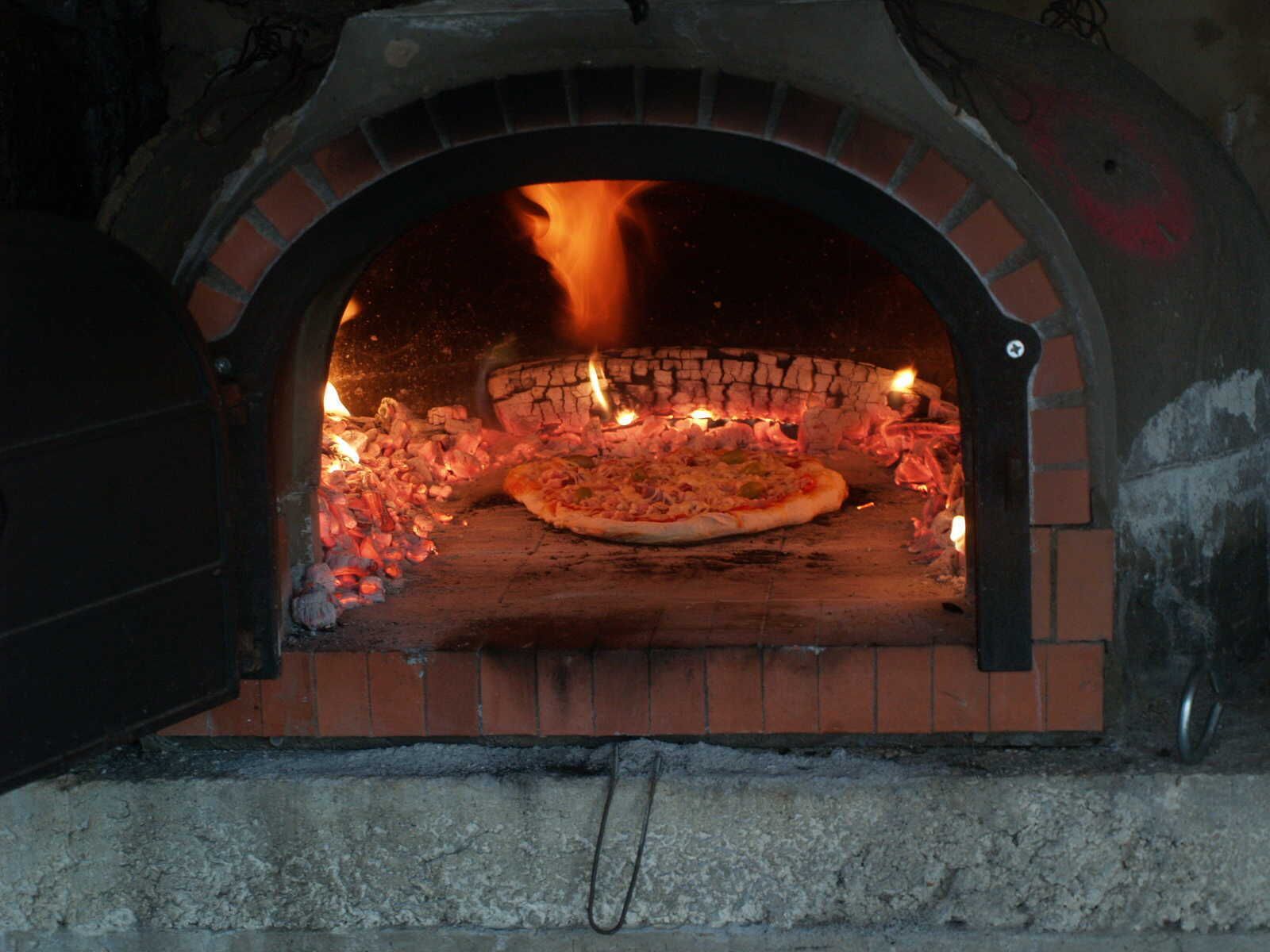 A brick oven pizza oven with a pizza inside, flames burning, and a partially open door.