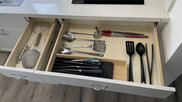 Un cassetto da cucina con utensili, un tagliere nero e un lavello in acciaio inossidabile.