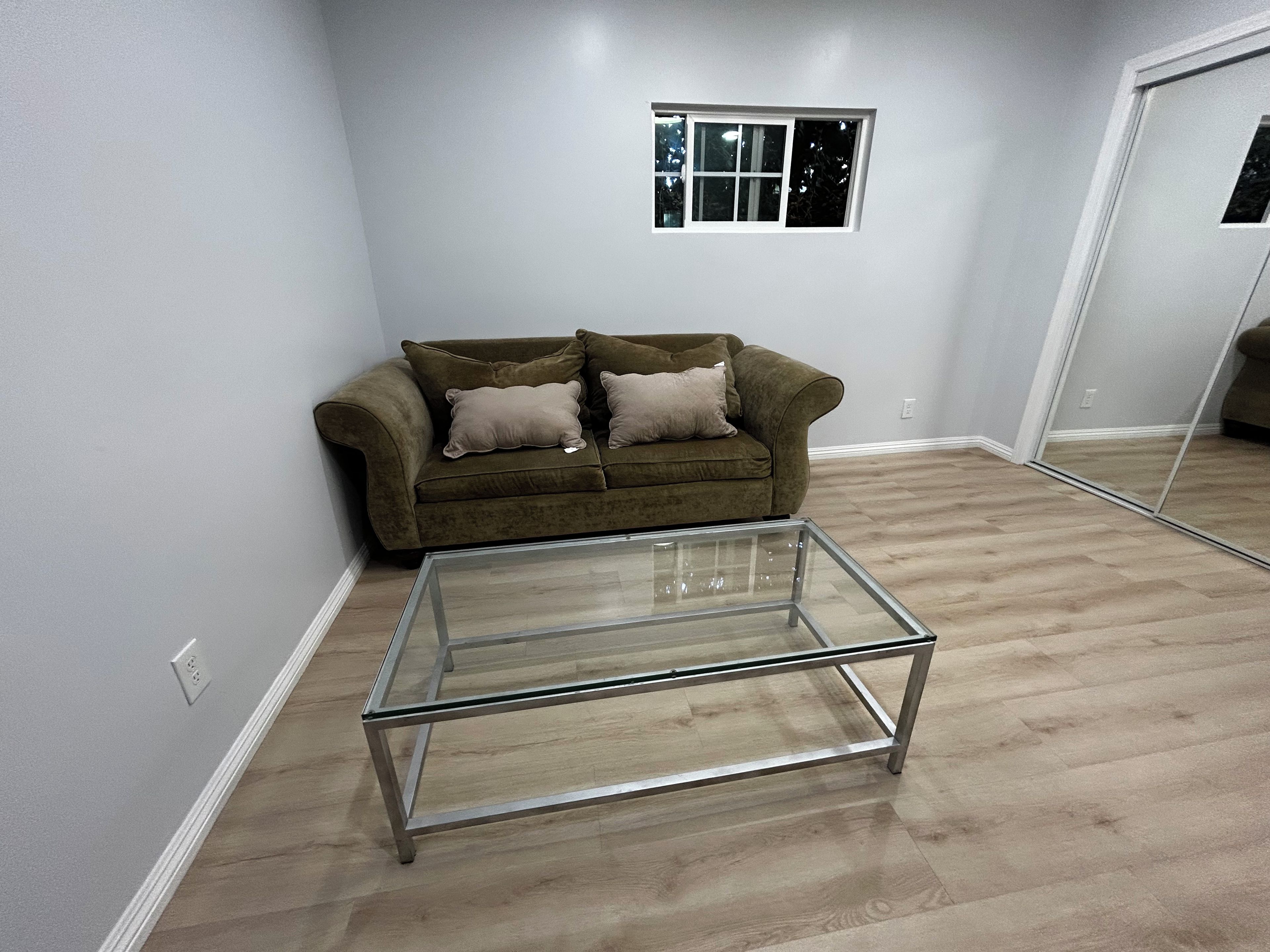 Una sala de estar con una mesa de centro de vidrio, un sofá marrón con cojines, un ventanal y un espejo en la pared.