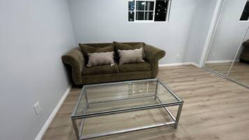 Una sala de estar con una mesa de centro de vidrio, un sofá marrón con cojines, un ventanal y un espejo en la pared.