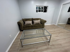 A living room with a glass coffee table, a brown sofa with cushions, a window, and a mirror on the wall.