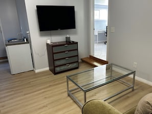 A modern living room with a glass coffee table, a flat-screen TV mounted on the wall, a small white refrigerator, and a wooden cabinet with drawers.