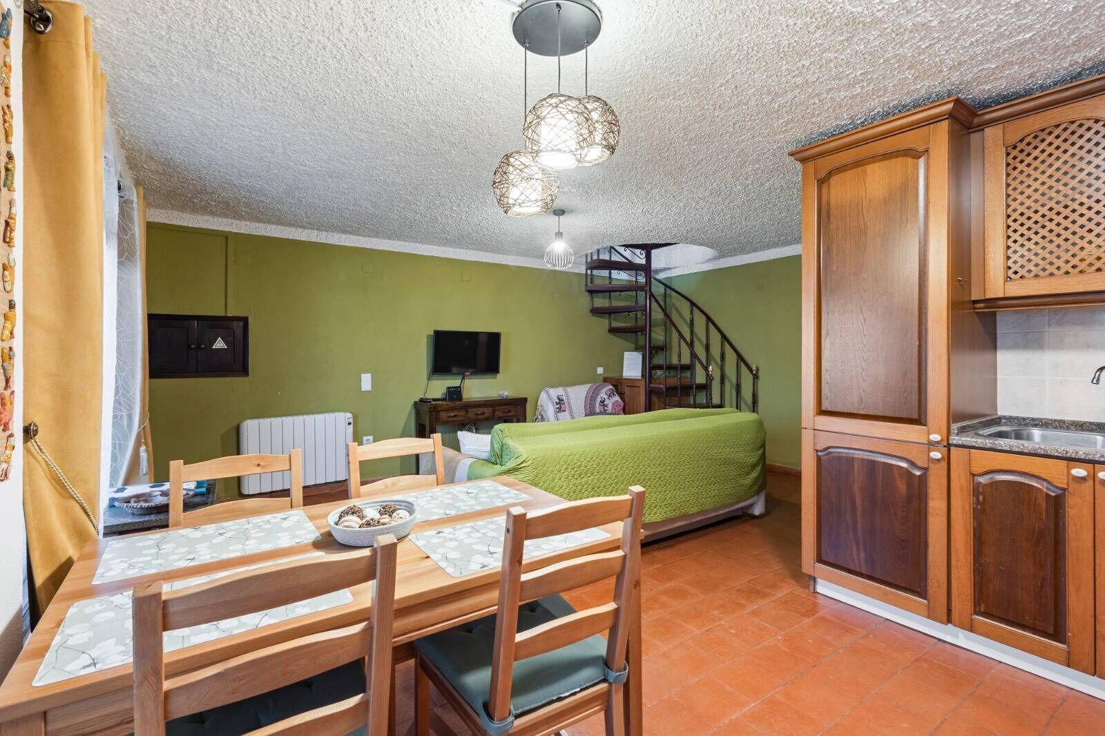 Une cuisine compacte avec des meubles en bois, une table à manger avec des chaises, un escalier et un lit recouvert d’une couverture verte.