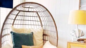 Ein runder Korbstuhl mit Kissen, ein kleiner Tisch mit einer Lampe und ein gerahmtes Bild auf einer Holzkommode.