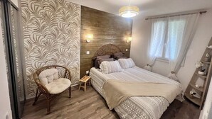 Ein Schlafzimmer mit Bett, Sessel, Nachttisch, Holz-Kopfteil und einem Fenster mit Vorhängen.