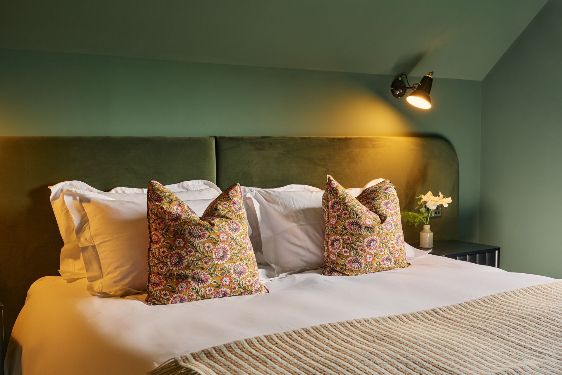 Ein ordentlich bezogenes Bett mit blumigen Kissen, ein Nachttisch mit Blumen und eine Wandlampe.
