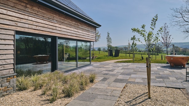 A modern building with a wooden facade, large glass windows, and a stone patio area. - Grain Loft - Luxury Cottages (Caersws)