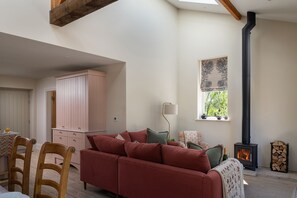 A cozy living room with a sofa, a table with chairs, a cabinet, a lamp, a window with a view, and a wood-burning stove. - Cherry Tree Cottage - Luxury Cottages (Lower Peover)