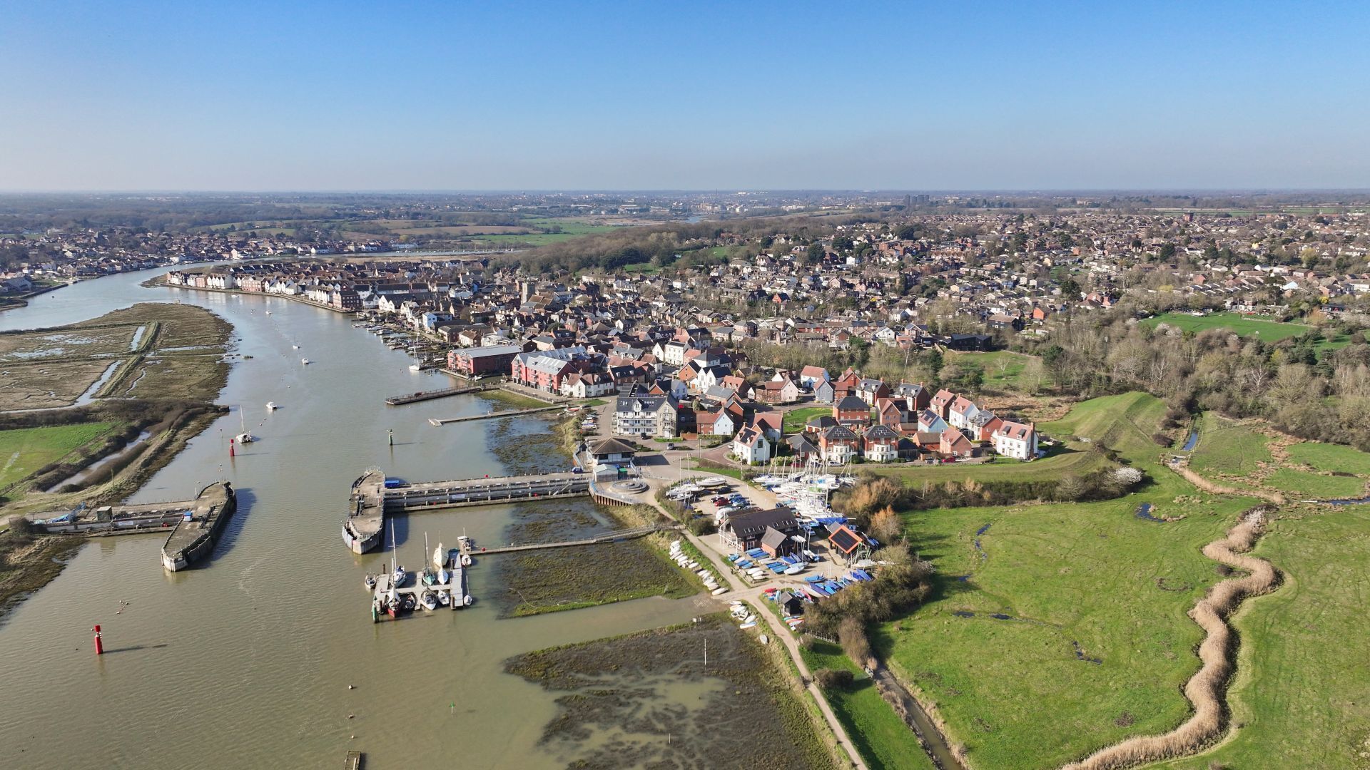 Vista aérea de un pueblo a orillas de un río, con un puerto deportivo, edificios residenciales y campos verdes.