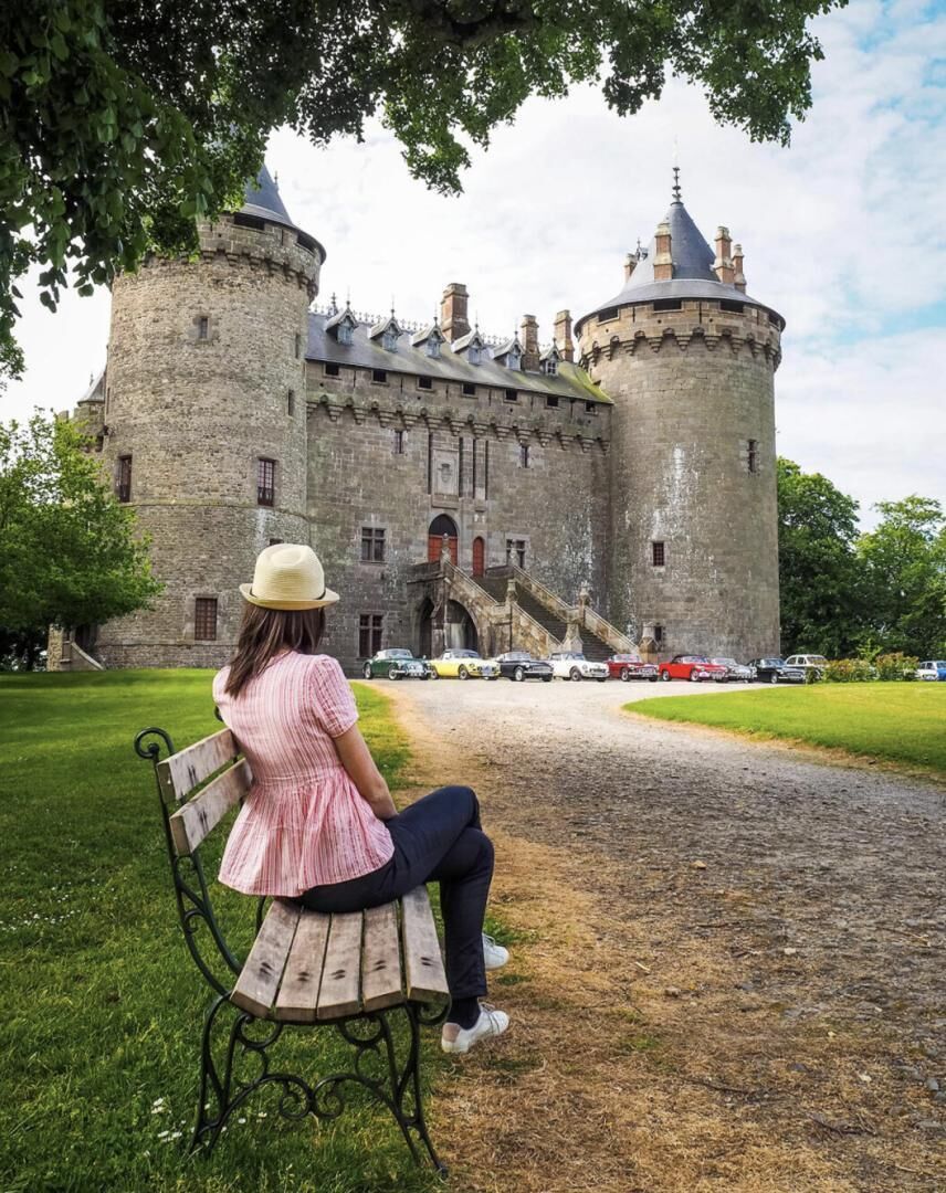 Mulher sentada num banco, de frente para um castelo com duas torres, um caminho calçado e carros estacionados.