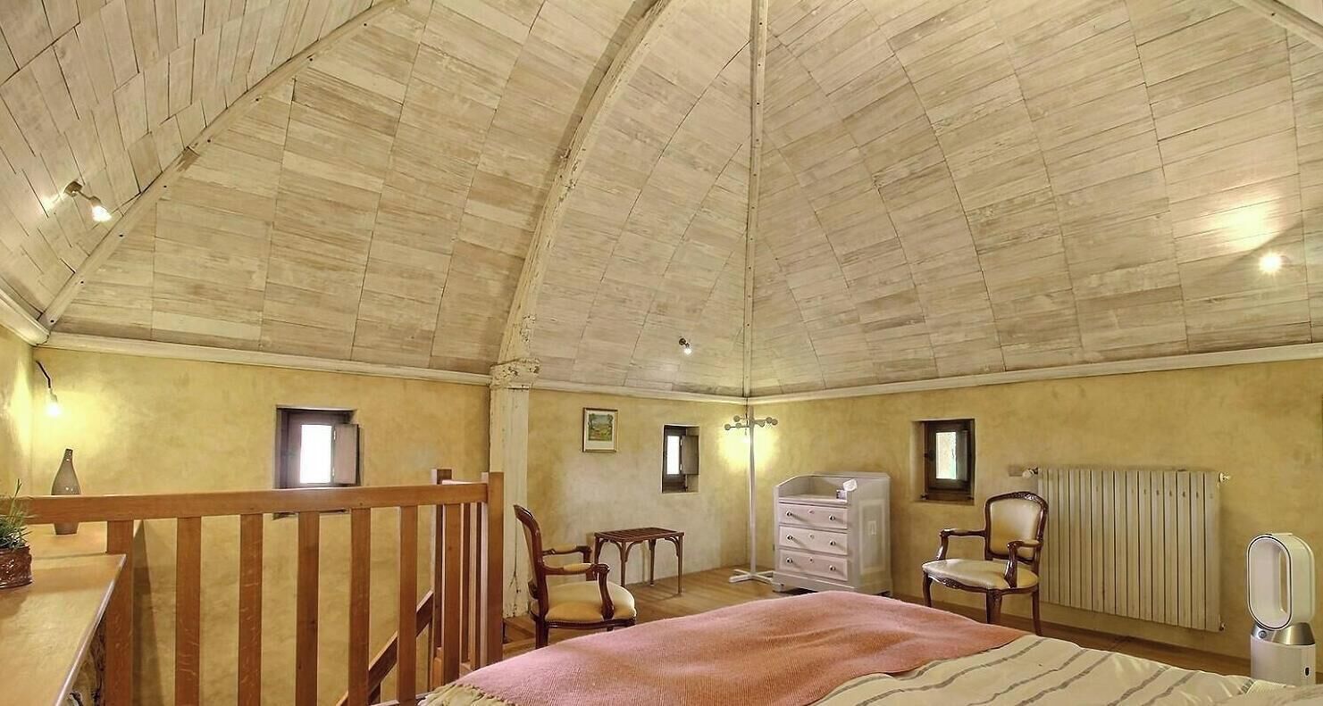 Ein geräumiges Schlafzimmer mit hoher Holzbalkendecke, einem Bett, einem Schminktisch, einem Sessel und einem Heizkörper.