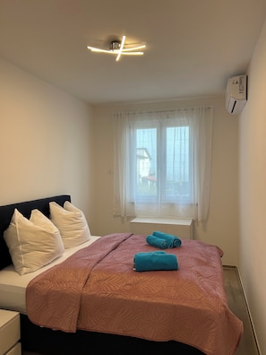 A bedroom with a bed, pillows, a window with curtains, a ceiling light fixture, and an air conditioning unit. - Luxurious new 3-room apartment with lake views and a pool on Lake Garda in Peschiera (Boschetti)