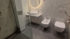 Ein modernes Badezimmer mit Duschkabine aus Glas, einem runden Spiegel, einem weißen Waschbecken, einem Bidet und einer Toilette.