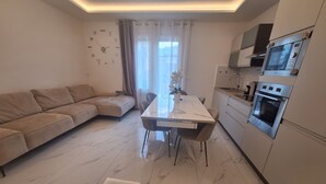 Ein zeitgemäßer Wohnbereich mit Sofa, Esstisch, Stühlen, Küchenzeile mit Geräte und einem fenster mit Vorhängen.