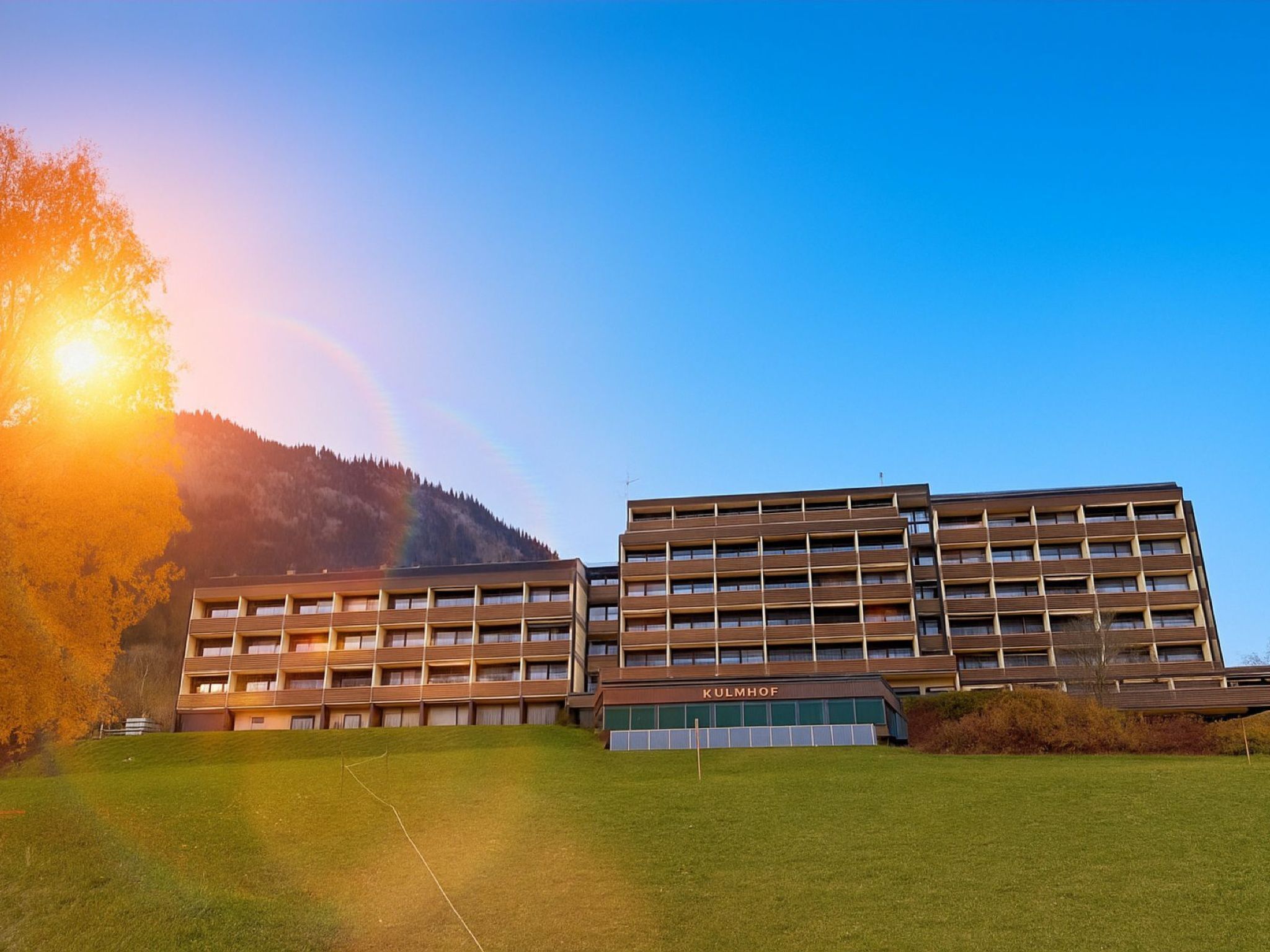 Un edificio moderno con tejado plano y varias ventanas, con la leyenda 'KULMHOF', ubicado en una colina cubierta de césped y cielo despejado.