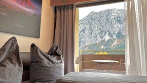 Et soveværelse med udsigt til et bjerg, en seng, et vindue med gardiner og et billede på væggen.