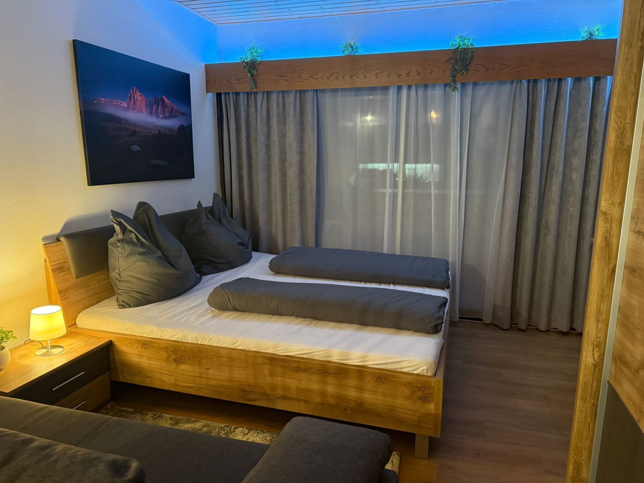 Een moderne slaapkamer met een houten bed, grijze beddengoed, een nachtkastje met een lamp en een schilderij aan de muur.