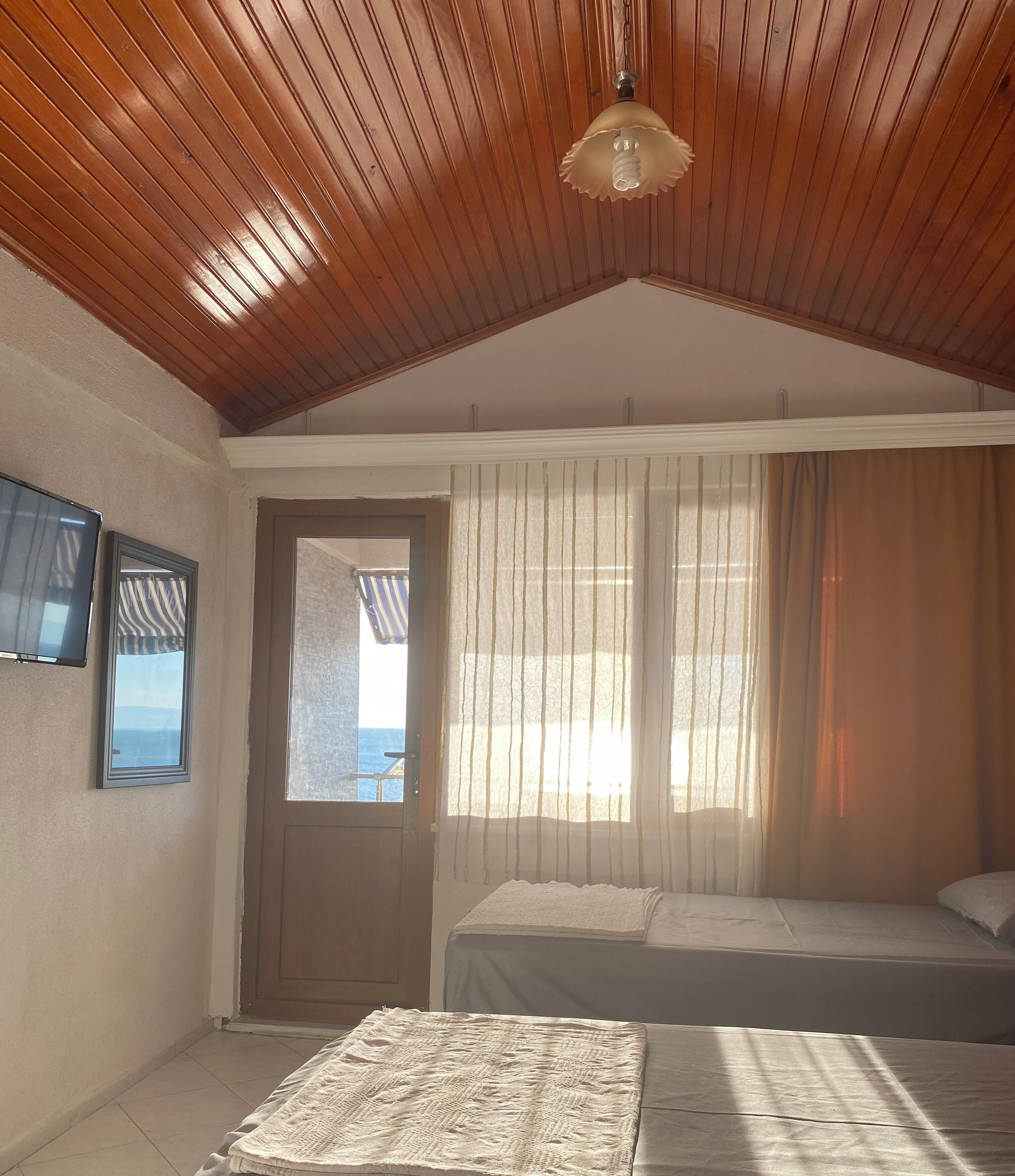 A bedroom with a wooden ceiling, a bed, a window with curtains, and a door.