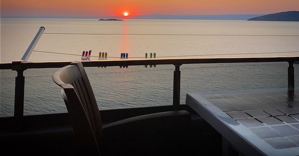 Vista desde un balcón sobre un mar tranquilo al atardecer, con una silla y una barandilla en primer plano.