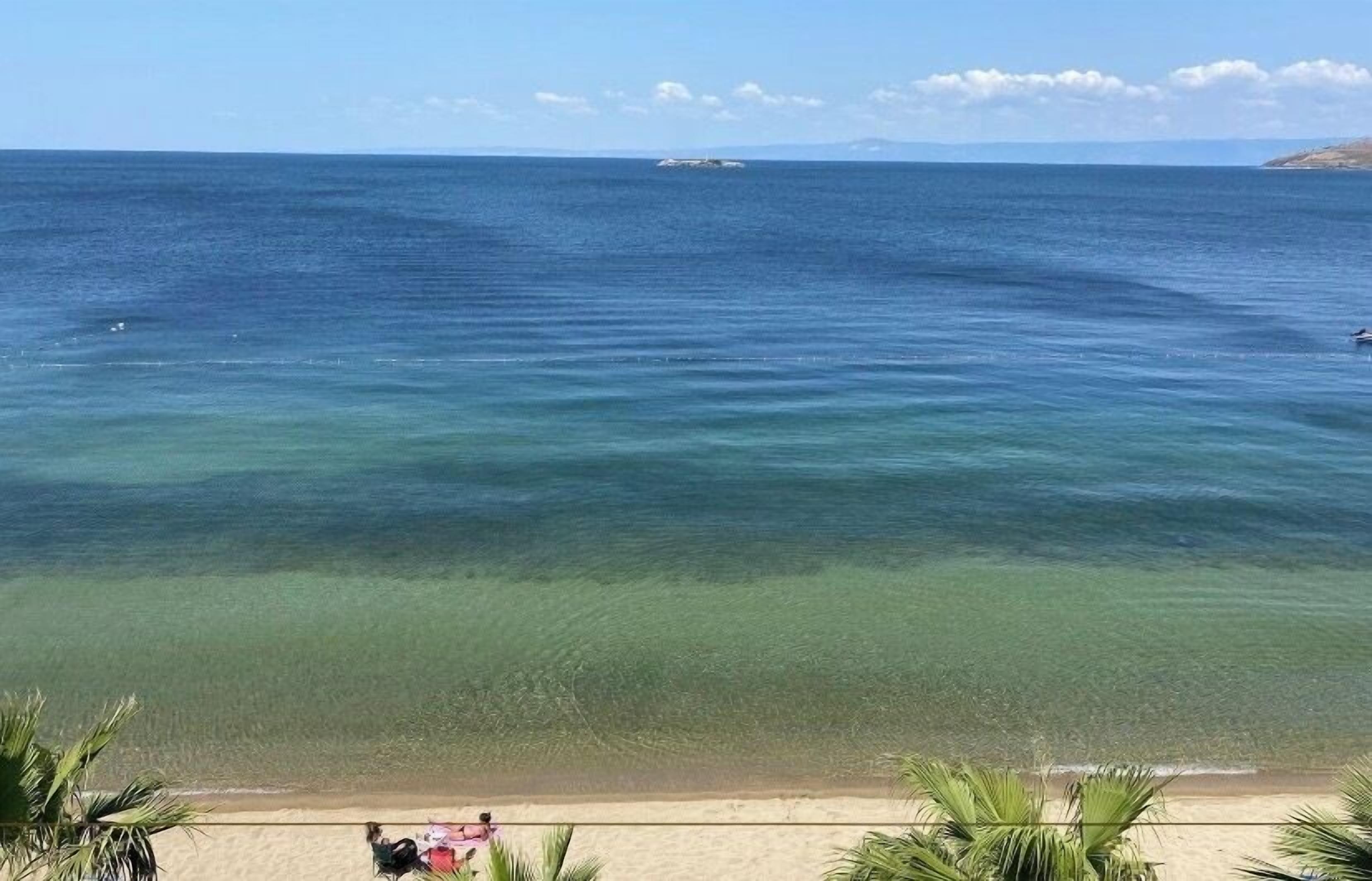 Una playa con agua cristalina, un barco a lo lejos y una persona sentada en la arena.