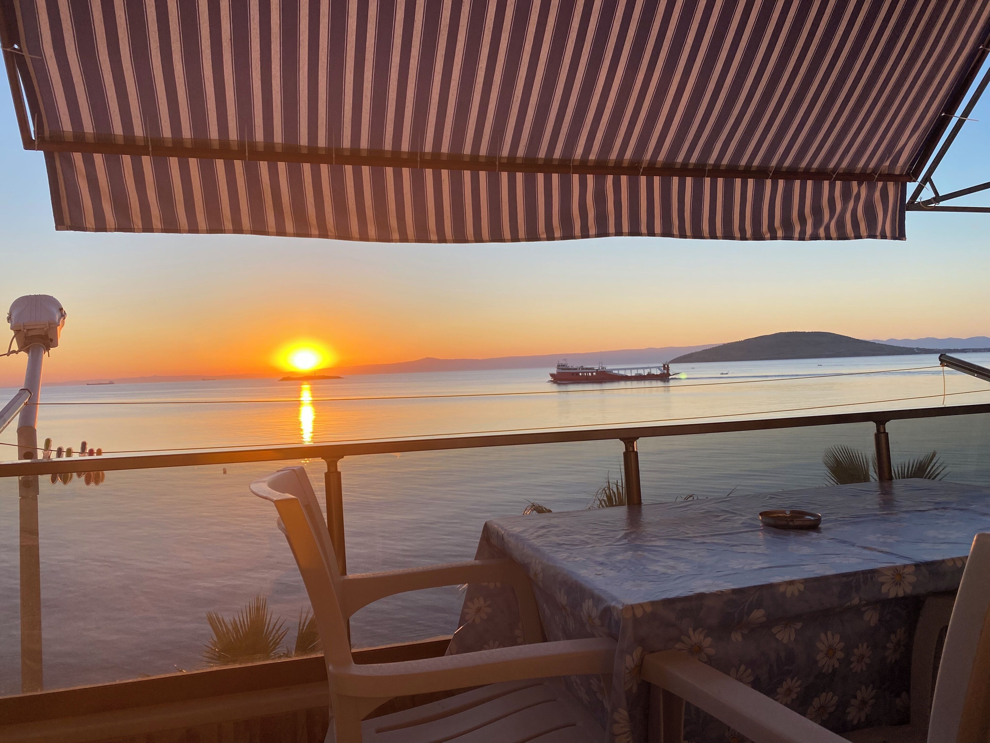 A terrace with a view of the sunset over the water, a table, chairs, and a railing.