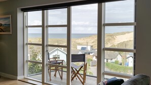 A room with large windows overlooking a beach and dunes, a folding chair, and a couch. - 4 The Rocks - sleeps 8 in 4 bedrooms (Holywell Bay)