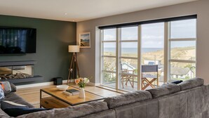 A modern living room with a large window, a flat-screen TV, a fireplace, and a cozy seating area. - 4 The Rocks - sleeps 8 in 4 bedrooms (Holywell Bay)