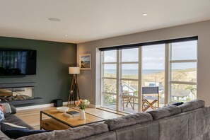 A modern living room with a large window, a flat-screen TV, a fireplace, and a cozy seating area. - 4 The Rocks - sleeps 8 in 4 bedrooms (Holywell Bay)