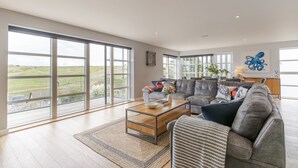 A spacious living room with large windows, a grey sofa, a coffee table, and a view of the outdoors. - 4 The Rocks - sleeps 8 in 4 bedrooms (Holywell Bay)
